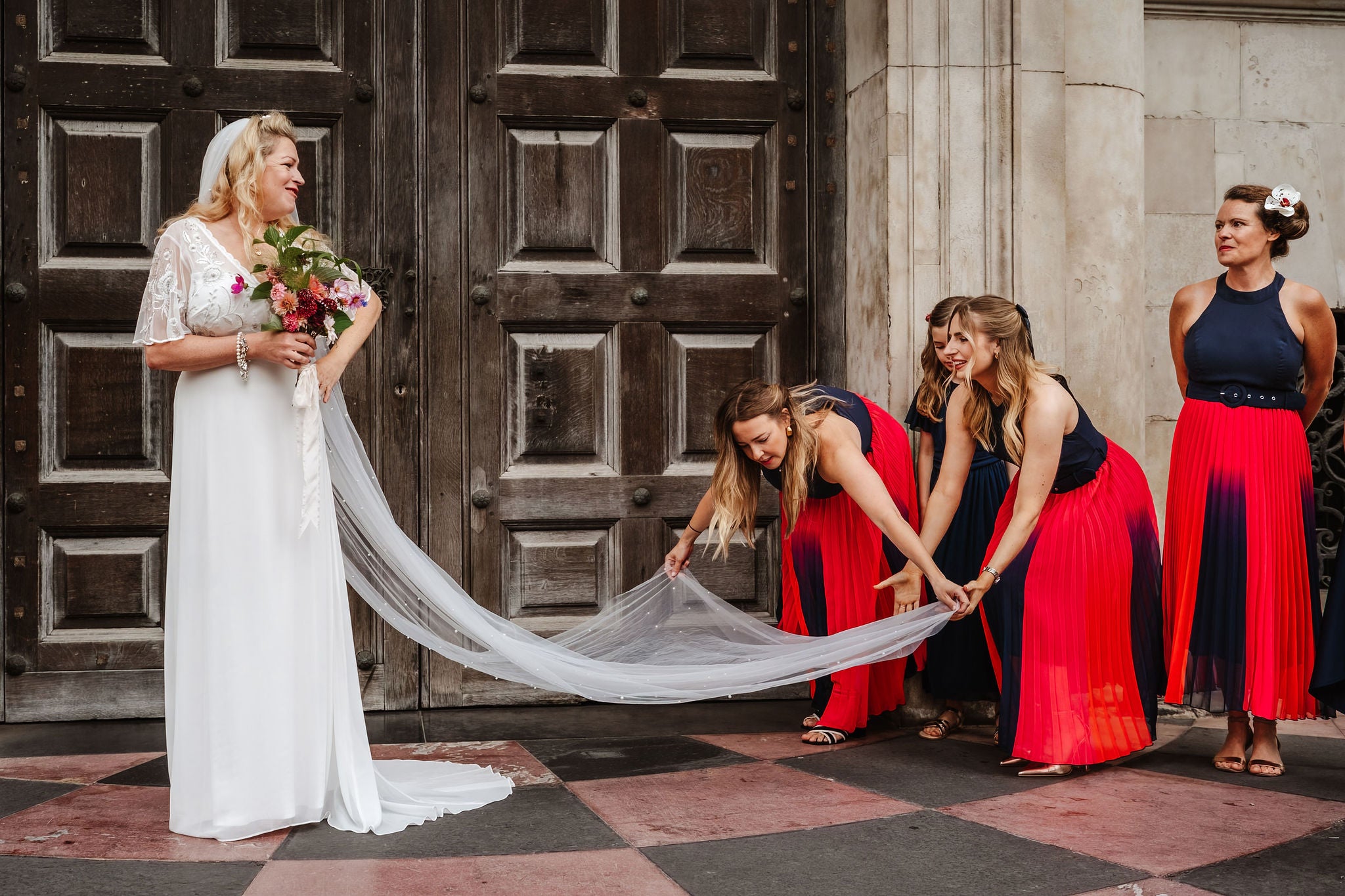 Ben Lee photograph of bride with beautiful bouquet, long veil and bridesmaids in red and black dresses standing in front of a large wooden door