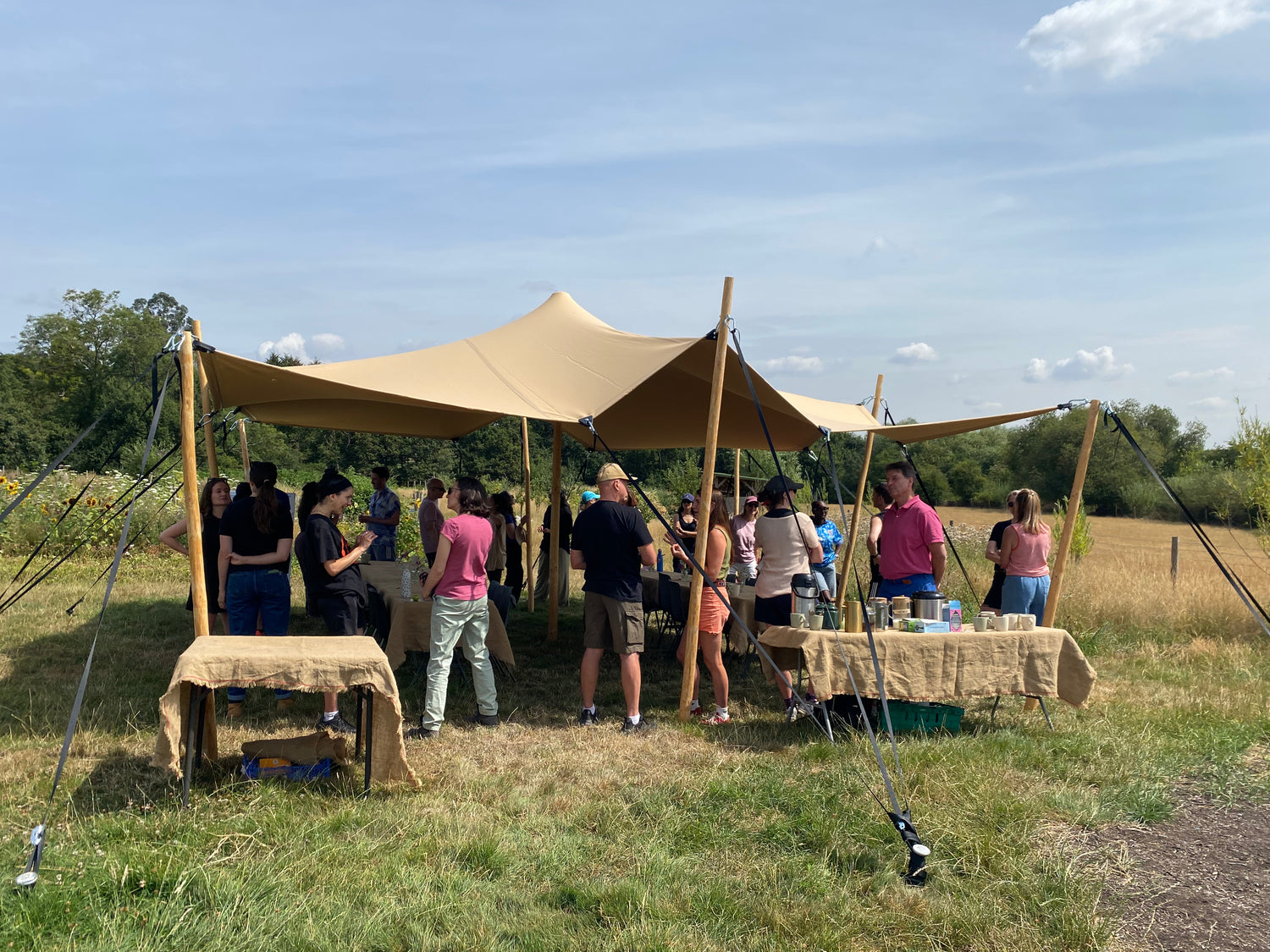 A large beige canvas event canopy set up outdoors in a grassy field, with several people standing and socialising underneath 